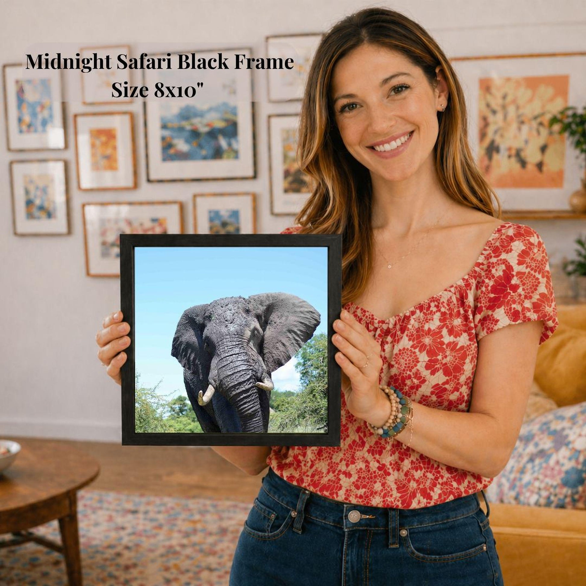 A woman is holding a framed picture of an elephant in a room with various framed pictures on the walls.