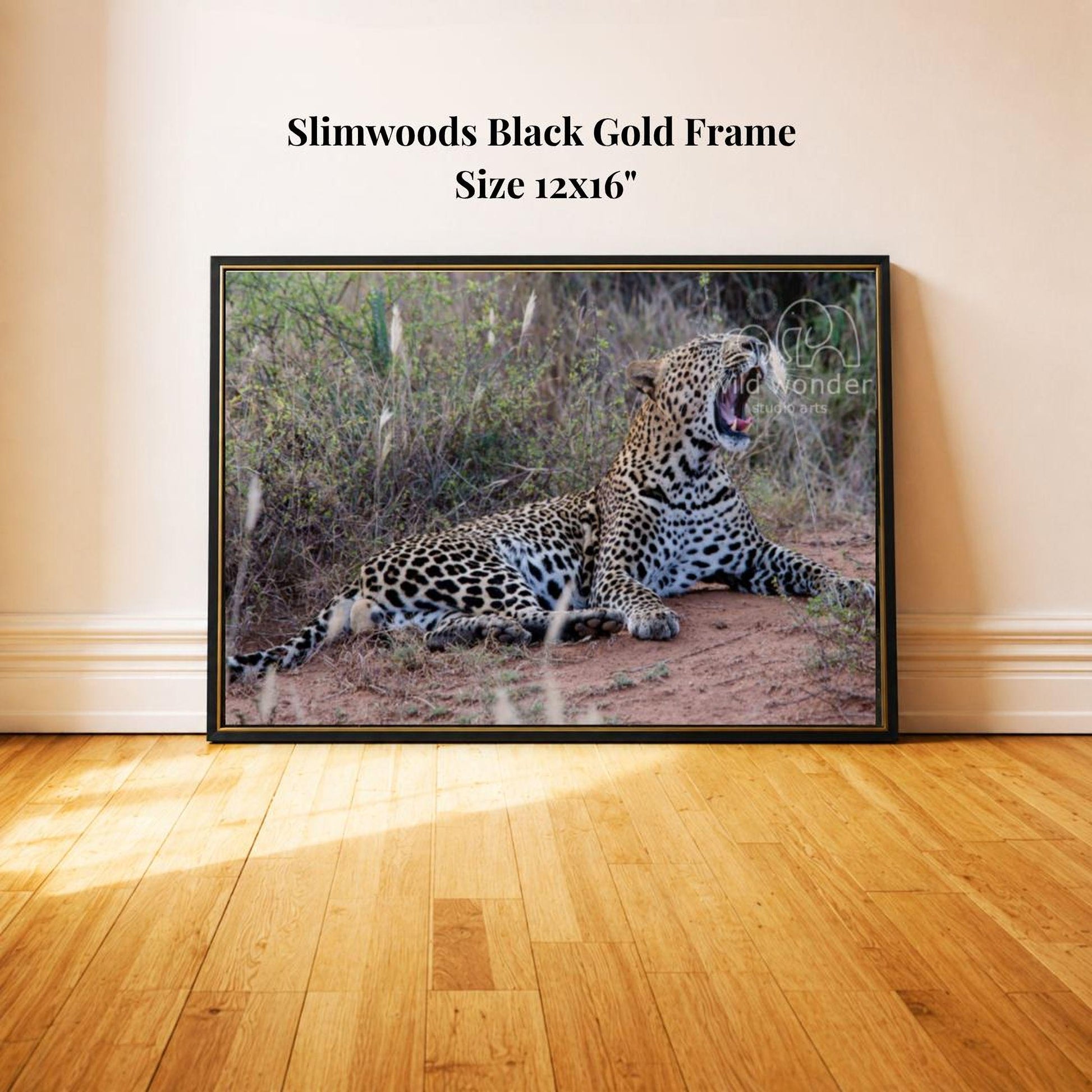 A large black framed picture of a leopard resting on the ground, with its mouth open, is displayed on a wooden floor against a white wall.