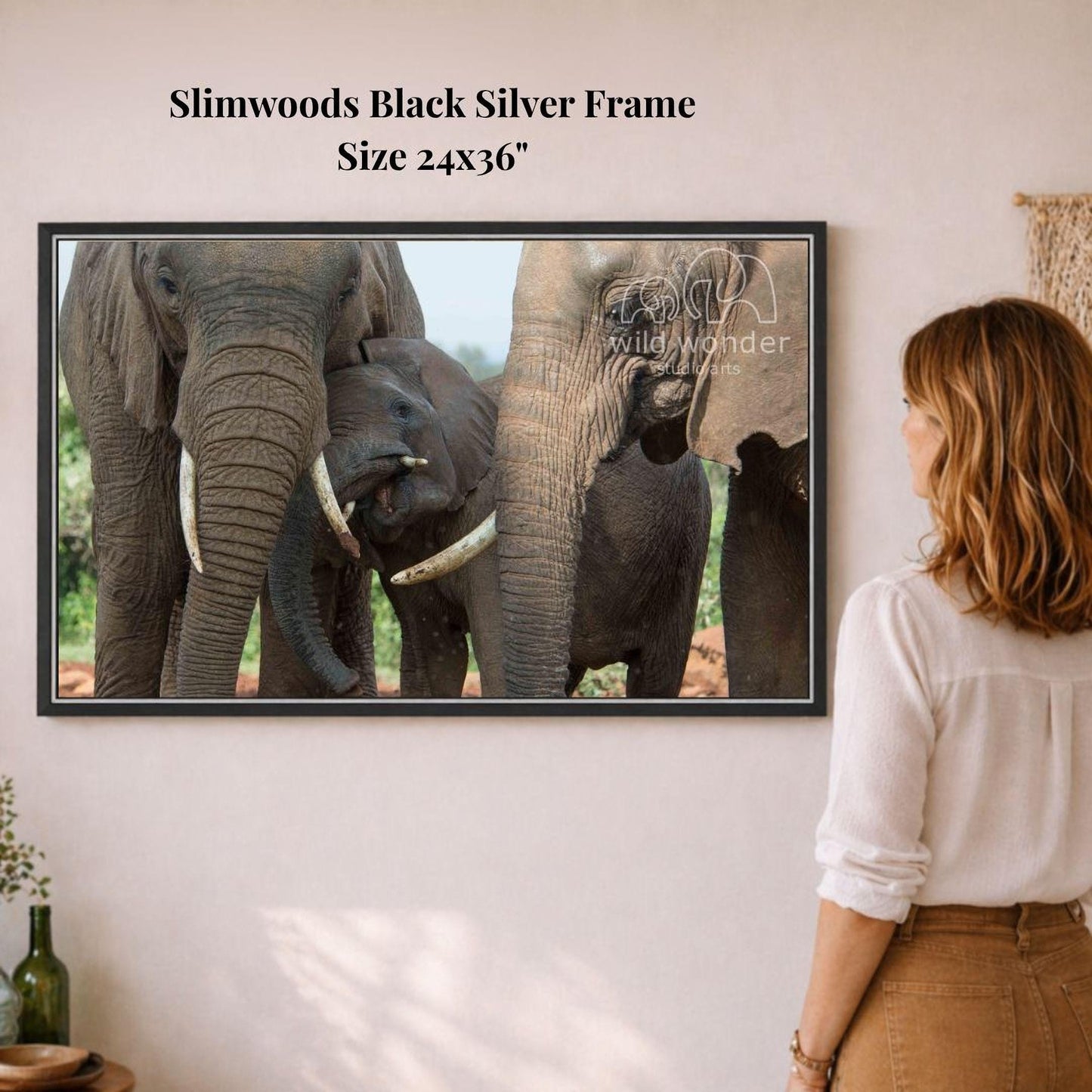A woman is standing in front of a framed photograph of three elephants, one of which is a baby elephant, on a wall.