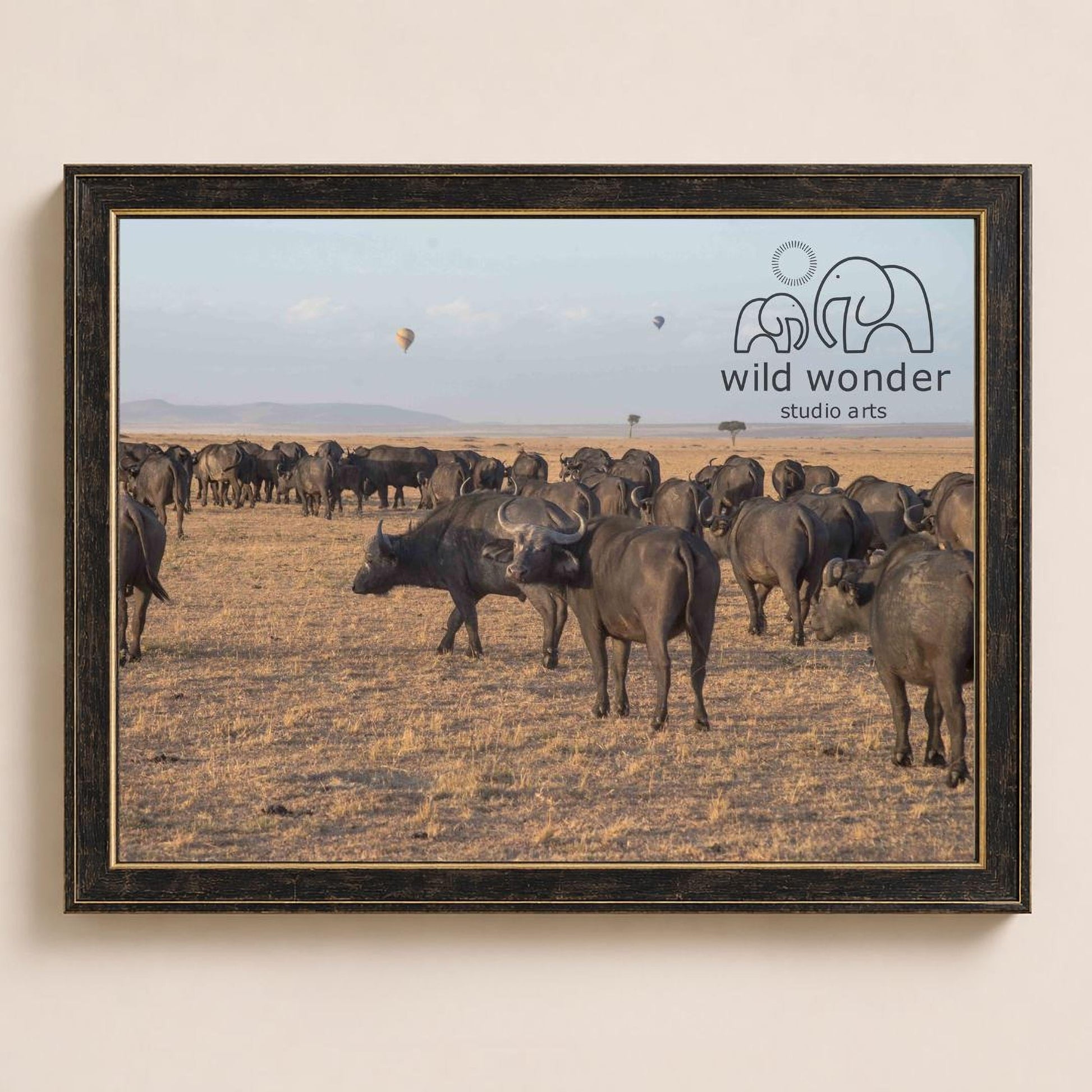 A herd of wild animals, including elephants and buffalo, grazing in a dry grassland landscape with a few hot air balloons visible in the sky.