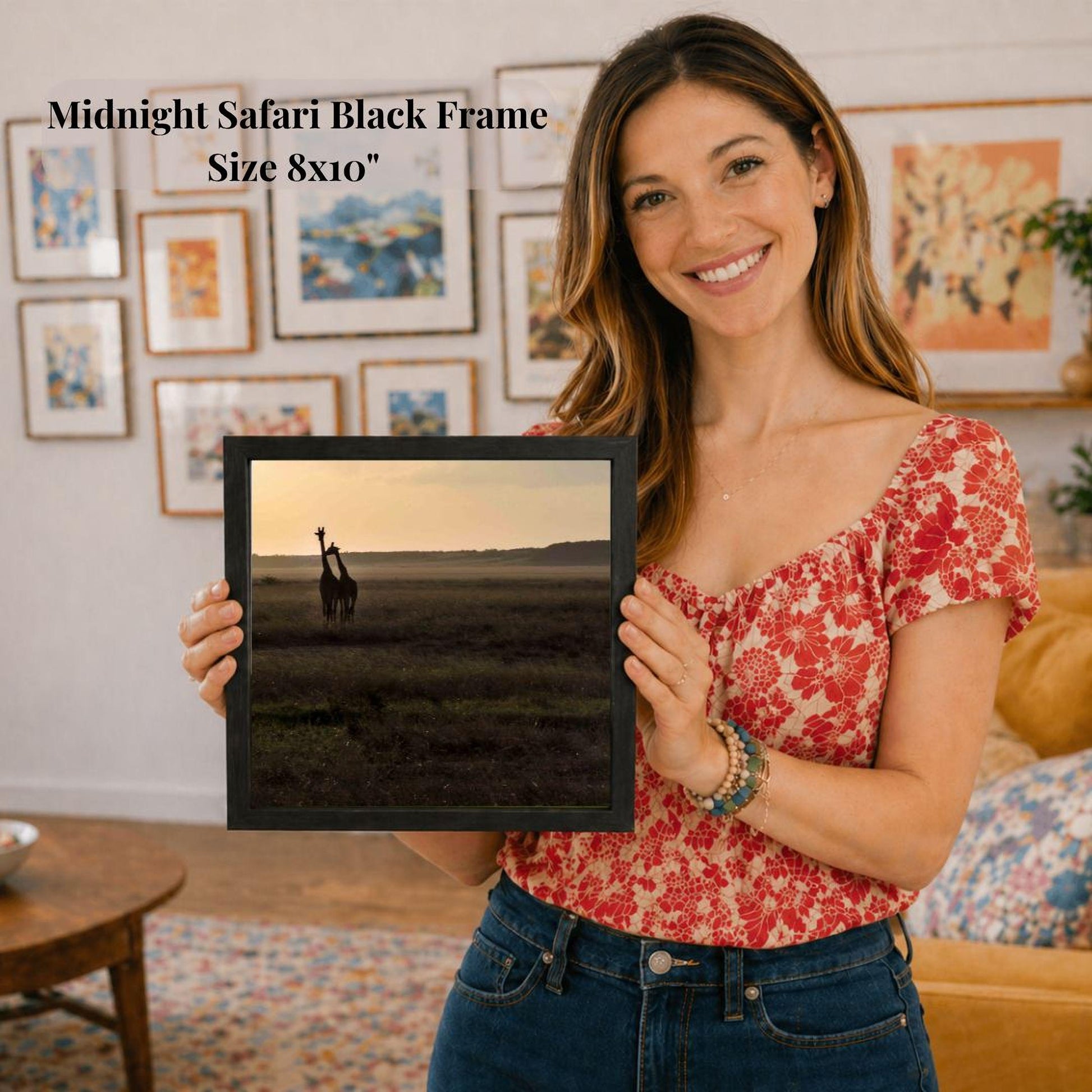 A woman is holding a framed photograph of two giraffes in a grassy field, with a smiling expression on her face.