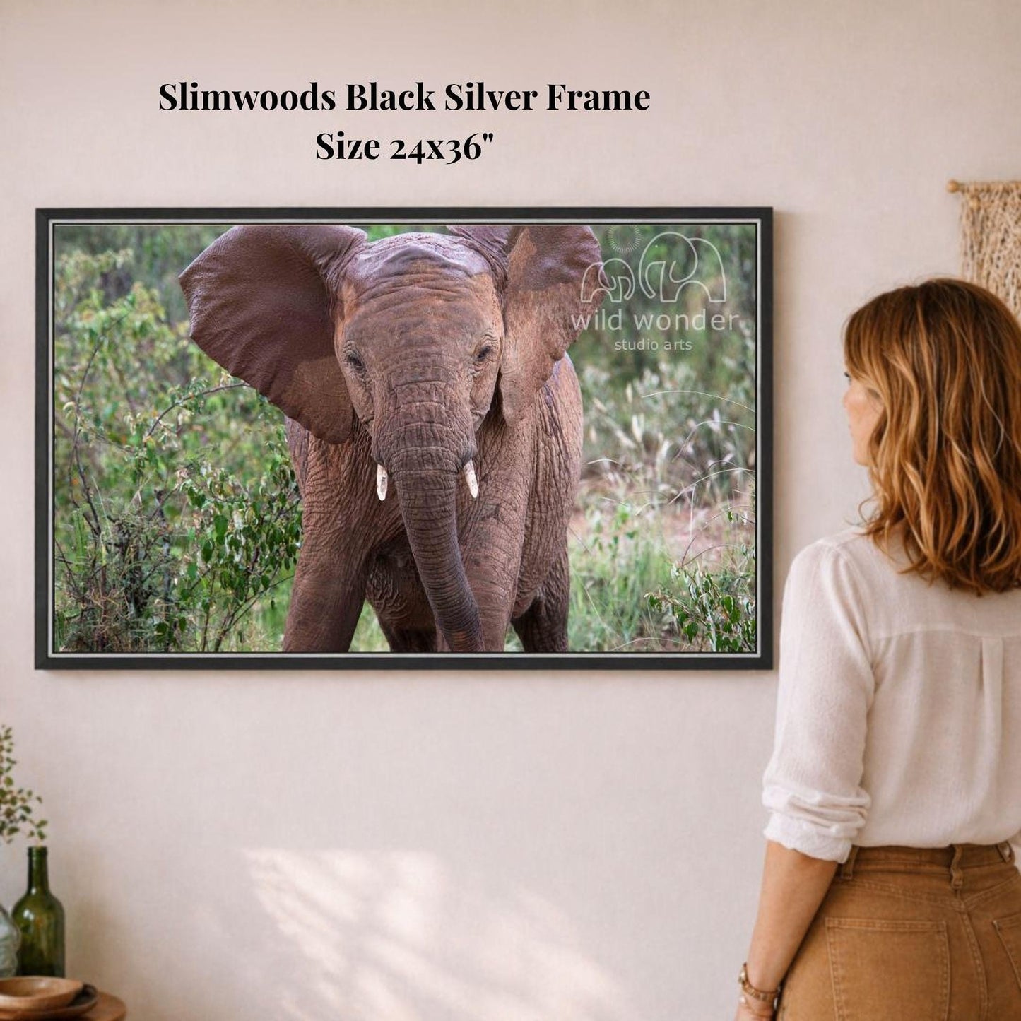 A woman is standing in front of a framed photograph of an elephant, observing the animal in a natural setting.