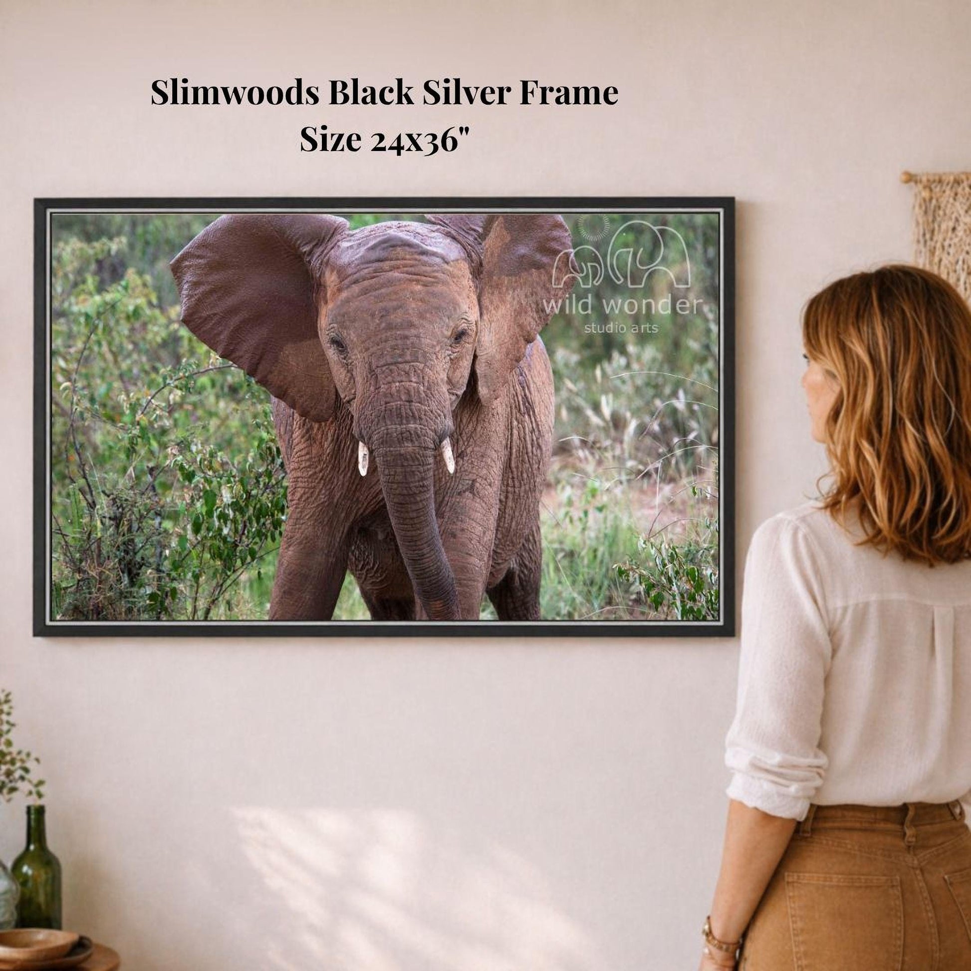 A woman is standing in front of a framed photograph of an elephant, observing the animal in a natural setting.