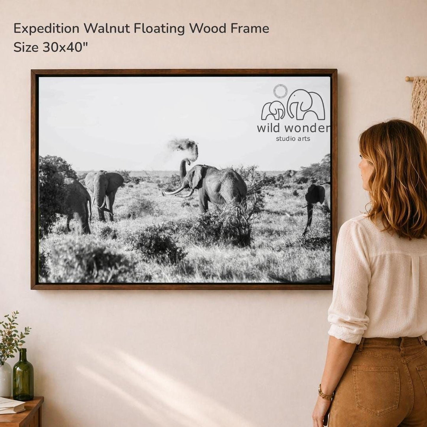 A woman is standing in front of a framed photograph of elephants in a field, looking at the artwork.
