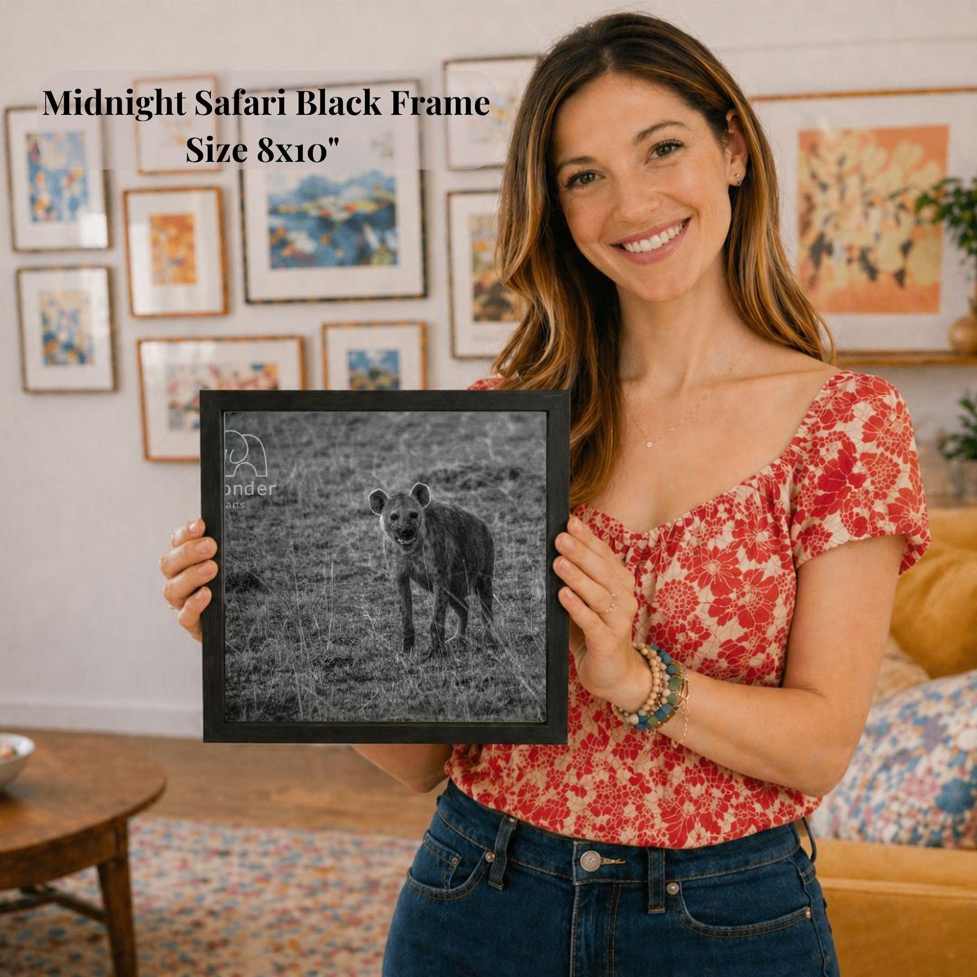 A woman is holding a framed photograph of a hyena in a grassy field, smiling and posing for the camera.
