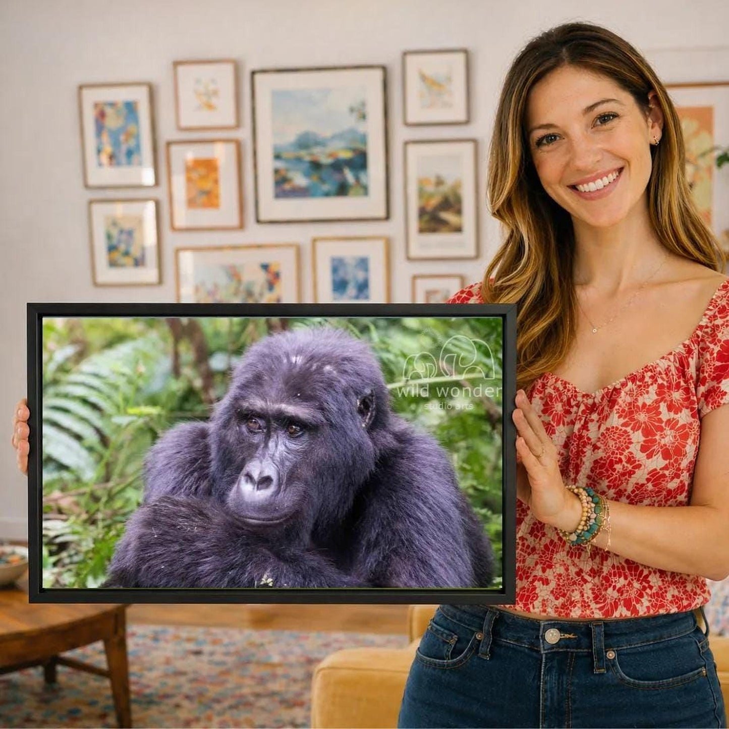 A woman is holding a framed photograph of a gorilla in front of a wall with various framed artworks.