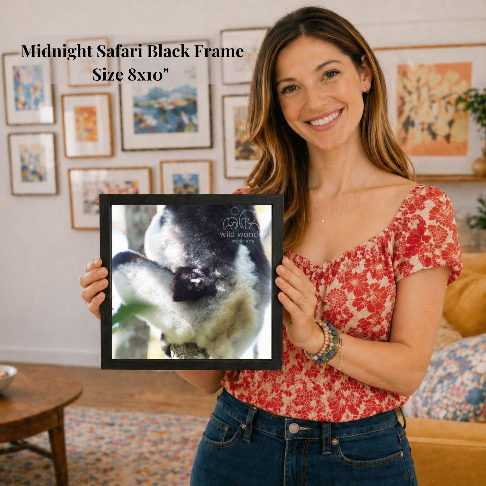 A woman is holding a framed photograph of a black and white animal, likely a cat, in front of a wall with various framed pictures.