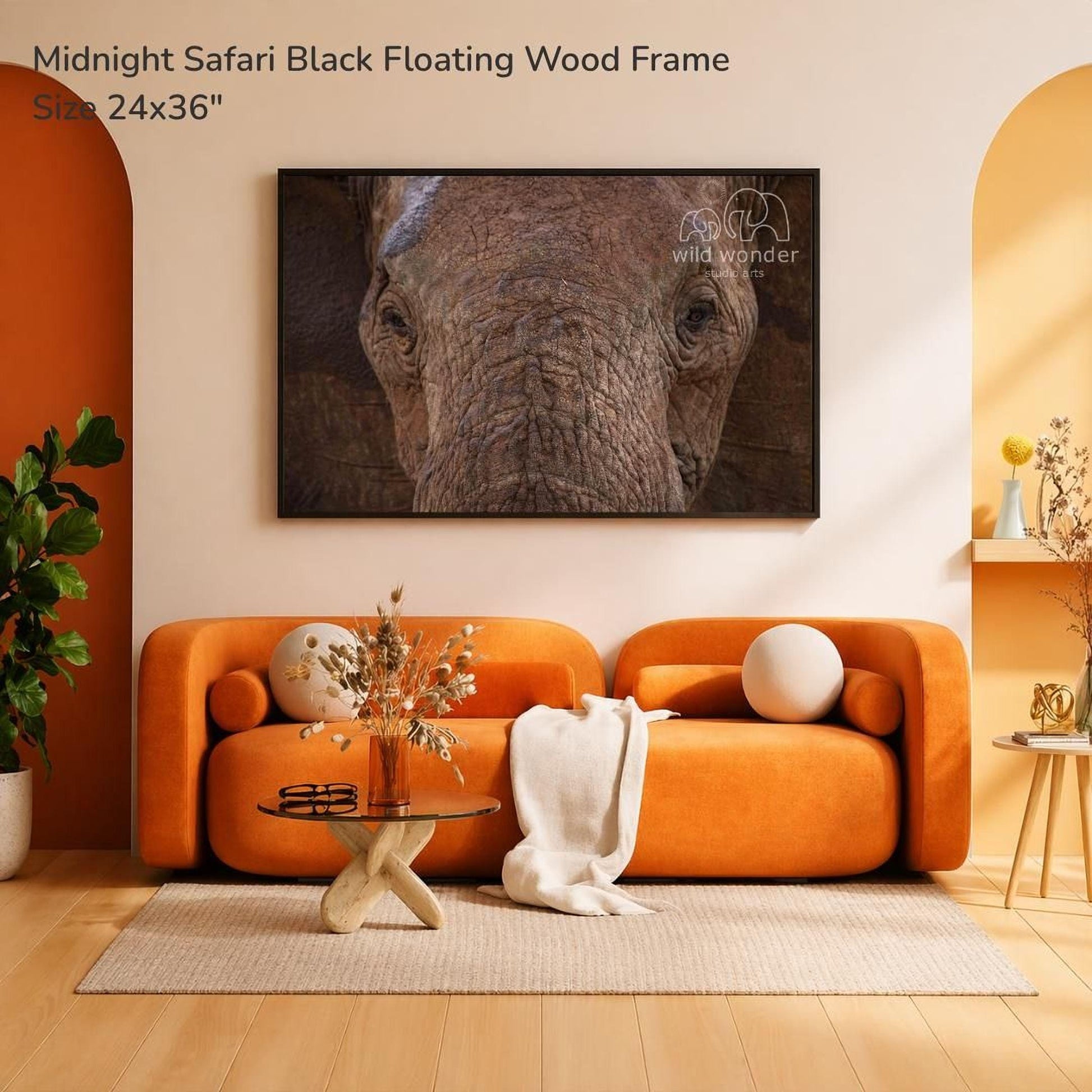a large black floating wood frame hanging on a wall, with a cozy living room setting below it. The living room has an orange sofa, a glass coffee table, and a potted plant. The overall scene has a warm and inviting atmosphere.