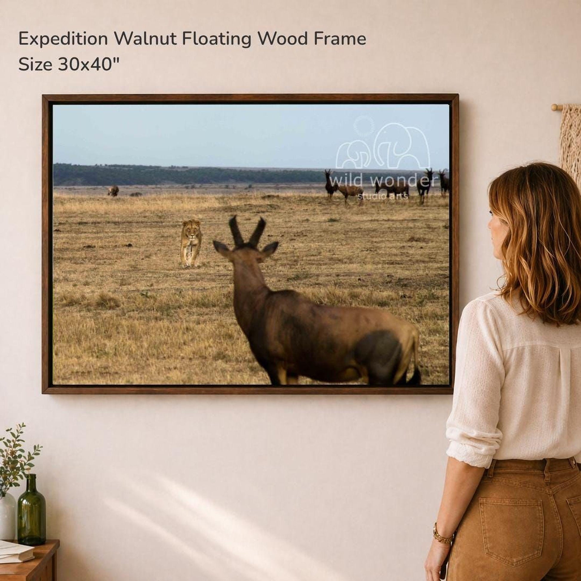 A woman is standing in front of a large framed photograph of a wild animal, such as a deer or a lion, in a grassy field.
