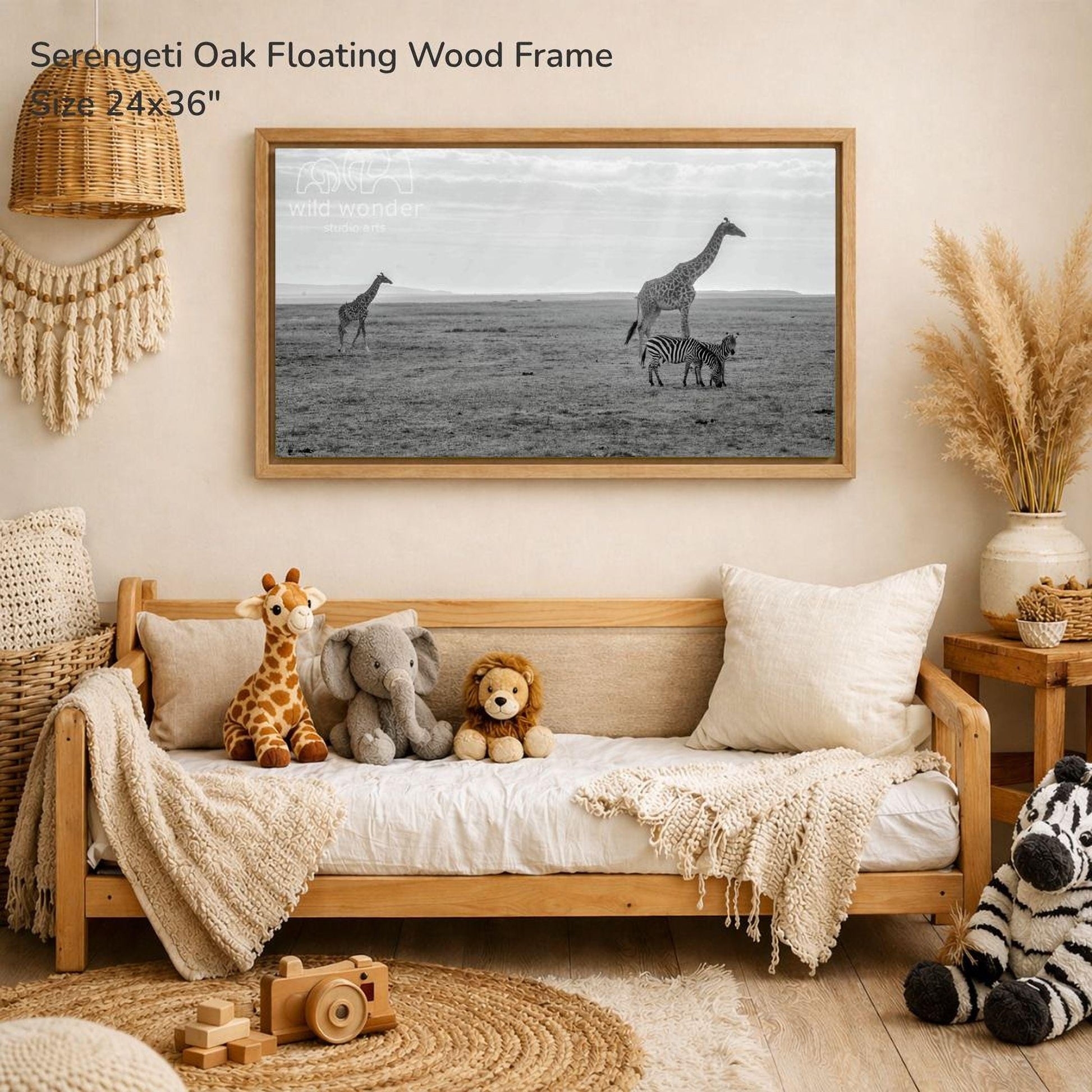 a serene indoor setting with a wooden frame hanging on a wall, displaying a black and white photograph of three giraffes in a field. The frame is described as a "Serengeti Oak Floating Wood Frame" and measures 24x36 inches. The room is decorated with various stuffed animals, including a giraffe, elephant, lion, and zebra, as well as a woven basket and a wooden toy car.
