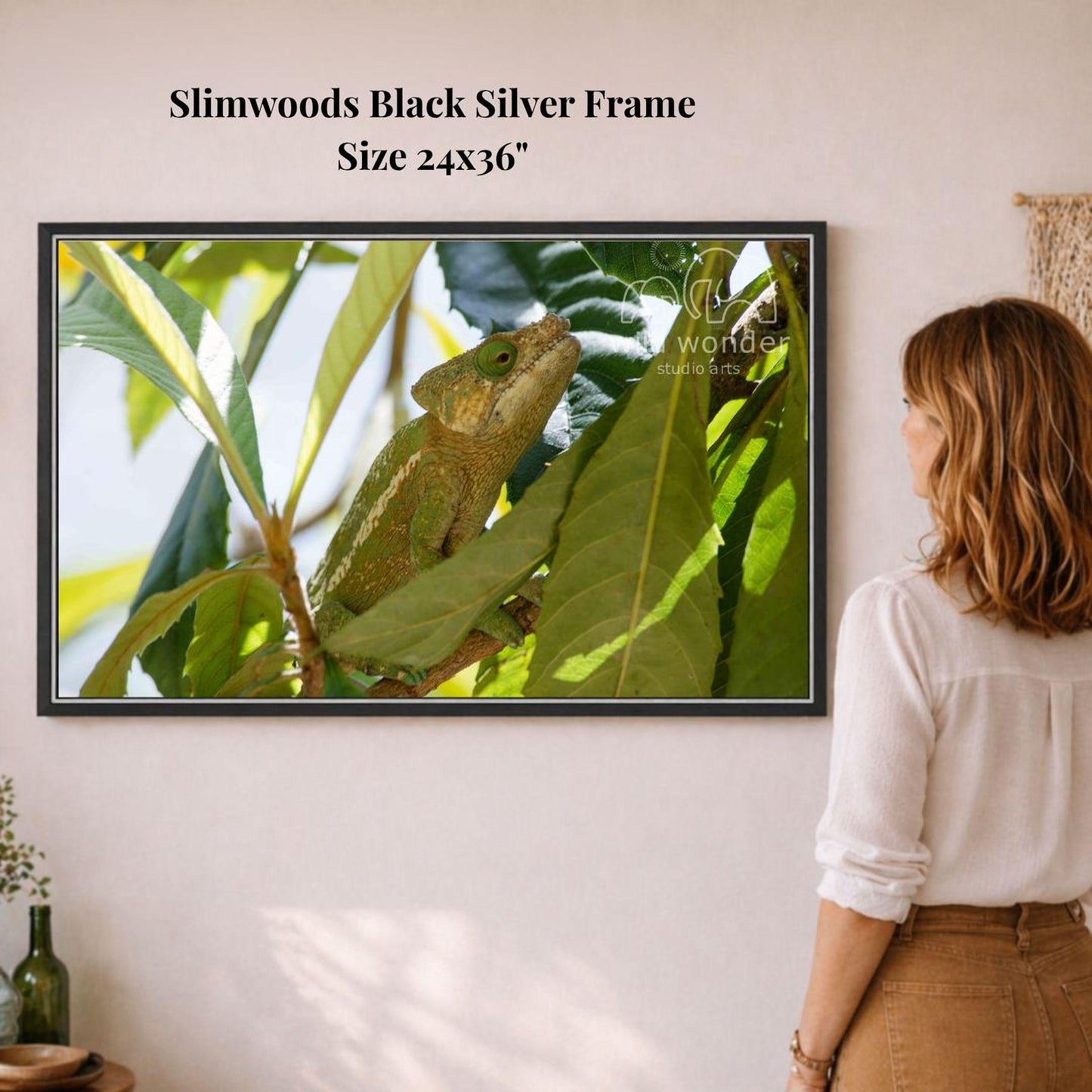 A woman is standing in front of a framed photograph of a chameleon perched on a leaf, which is hanging on a wall.