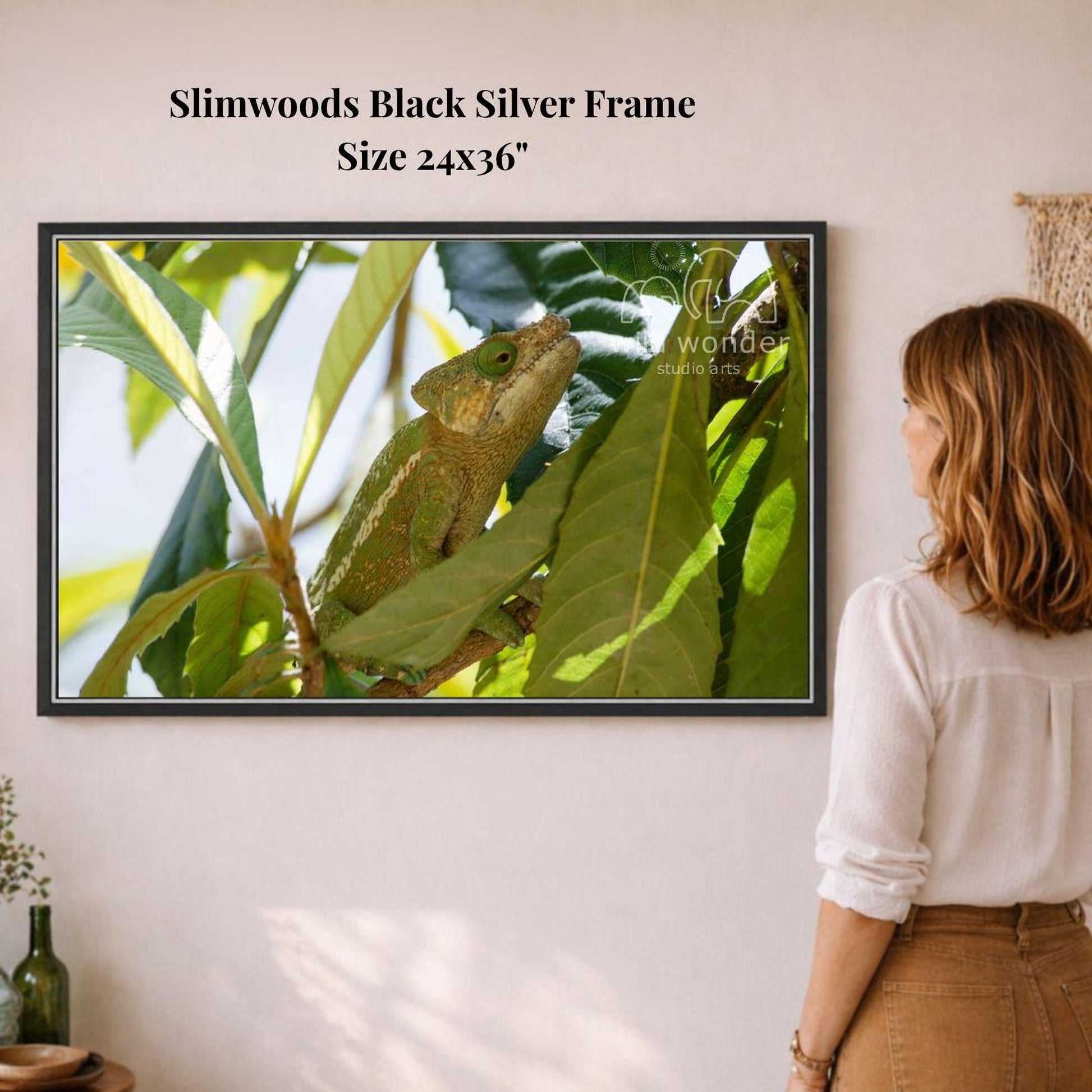 A woman is standing in front of a framed photograph of a chameleon perched on a leaf, which is hanging on a wall.