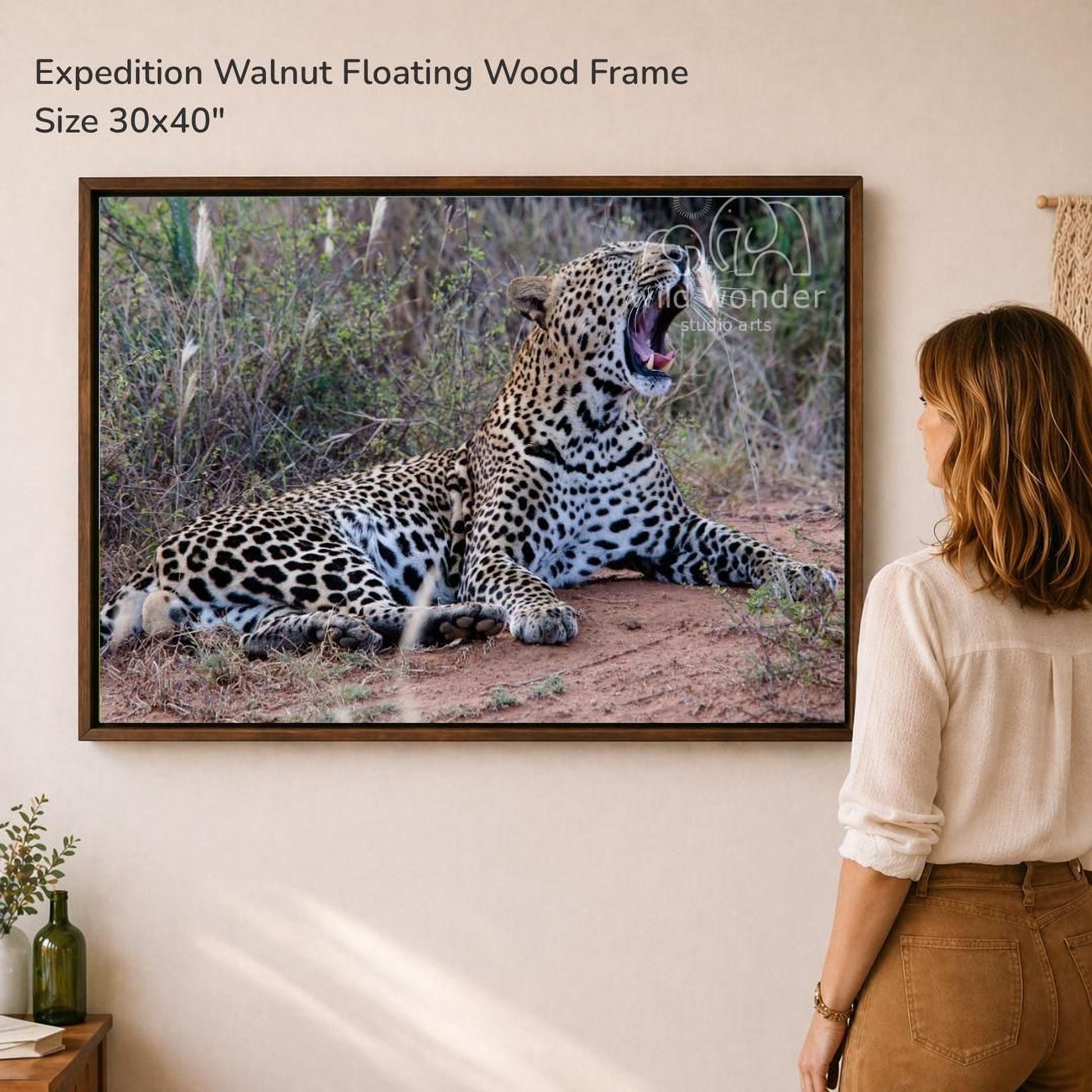 A woman is standing in front of a framed photograph of a leopard, which is lying on the ground in a natural setting.