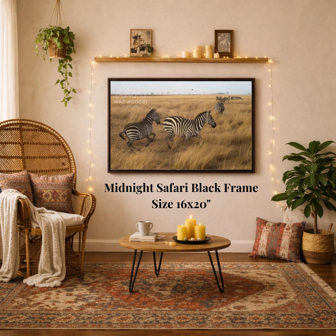 a living room with a large framed photograph of two zebras in a grassy field hanging on the wall. The photograph is in a black frame and is 16x20 inches in size. The room is decorated with a wicker chair, a coffee table, and a potted plant. The overall setting appears to be a cozy and comfortable living space.