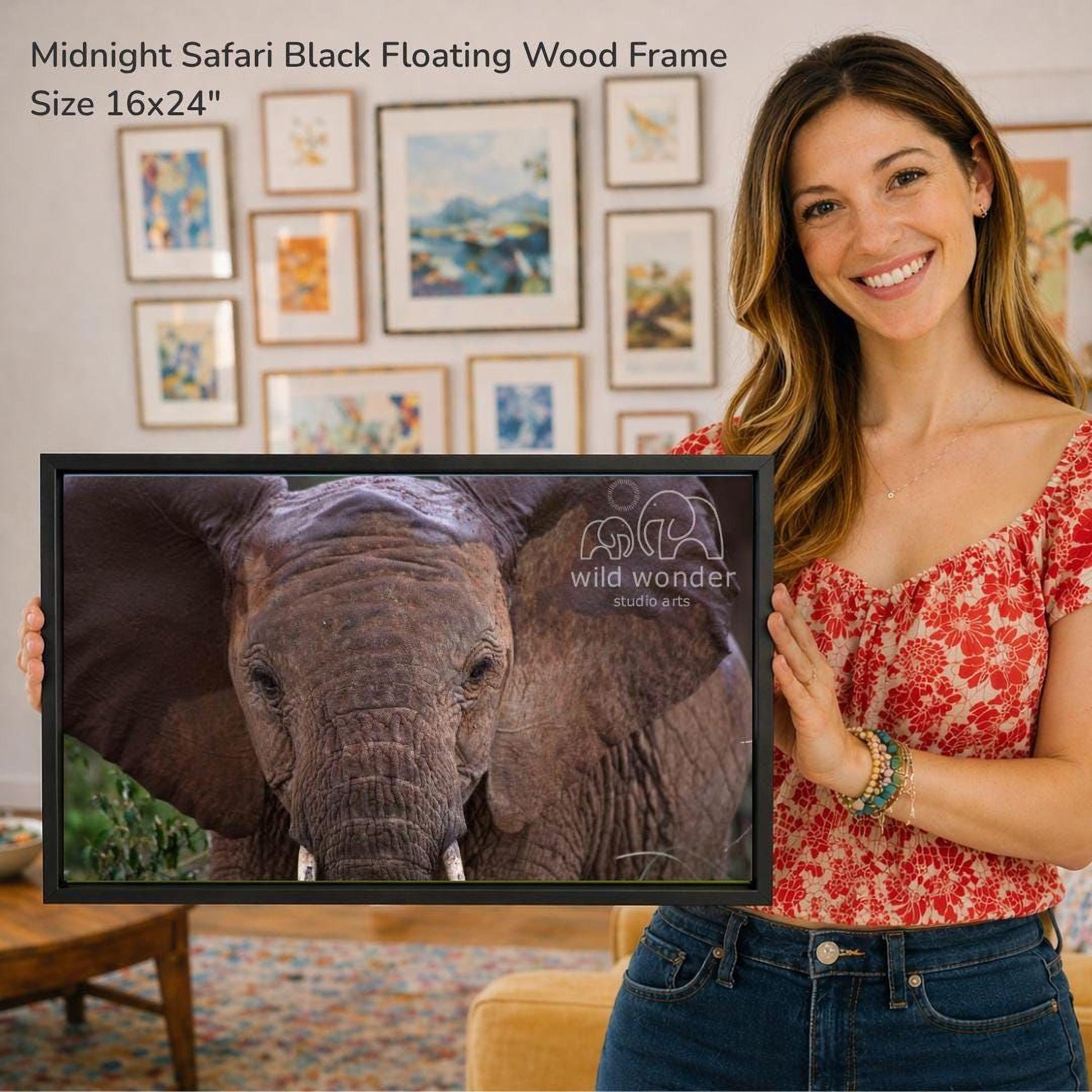 A woman is holding a framed photograph of an elephant in front of a wall with various framed artworks.
