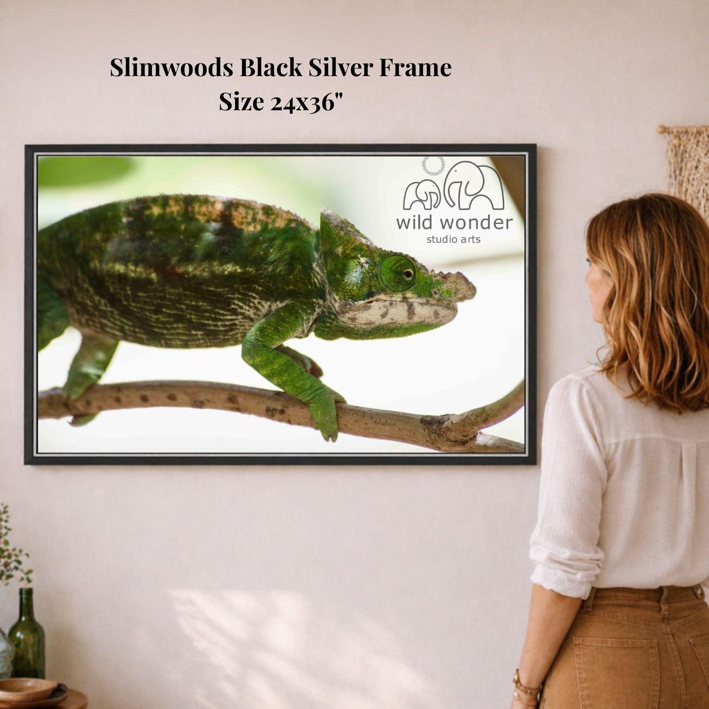 A woman is standing in front of a framed photograph of a green chameleon on a branch, looking at the artwork.