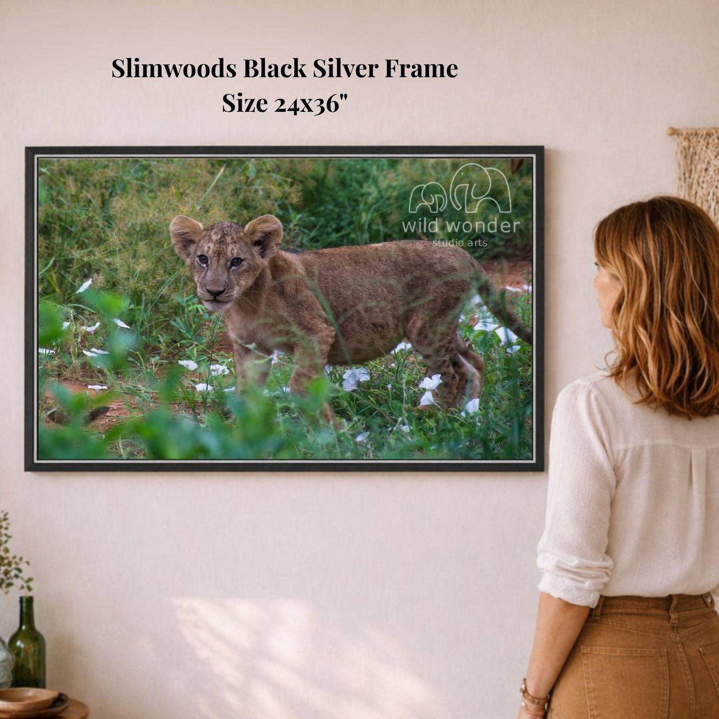 A woman is standing in front of a framed photograph of a young lion in a grassy field, looking at the artwork.