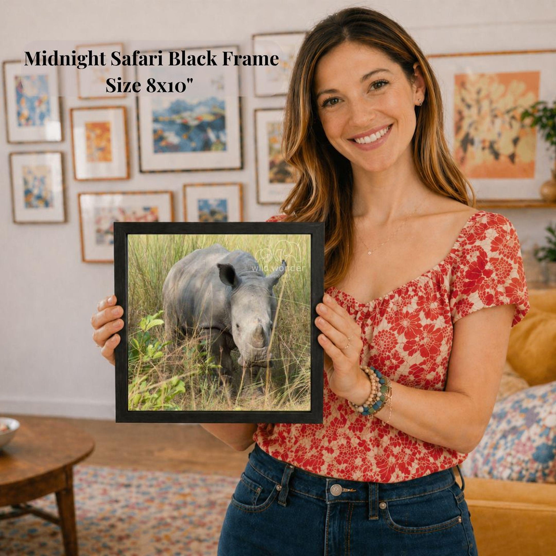 A woman is holding a framed photograph of a rhinoceros in a grassy field, smiling and posing for the camera.