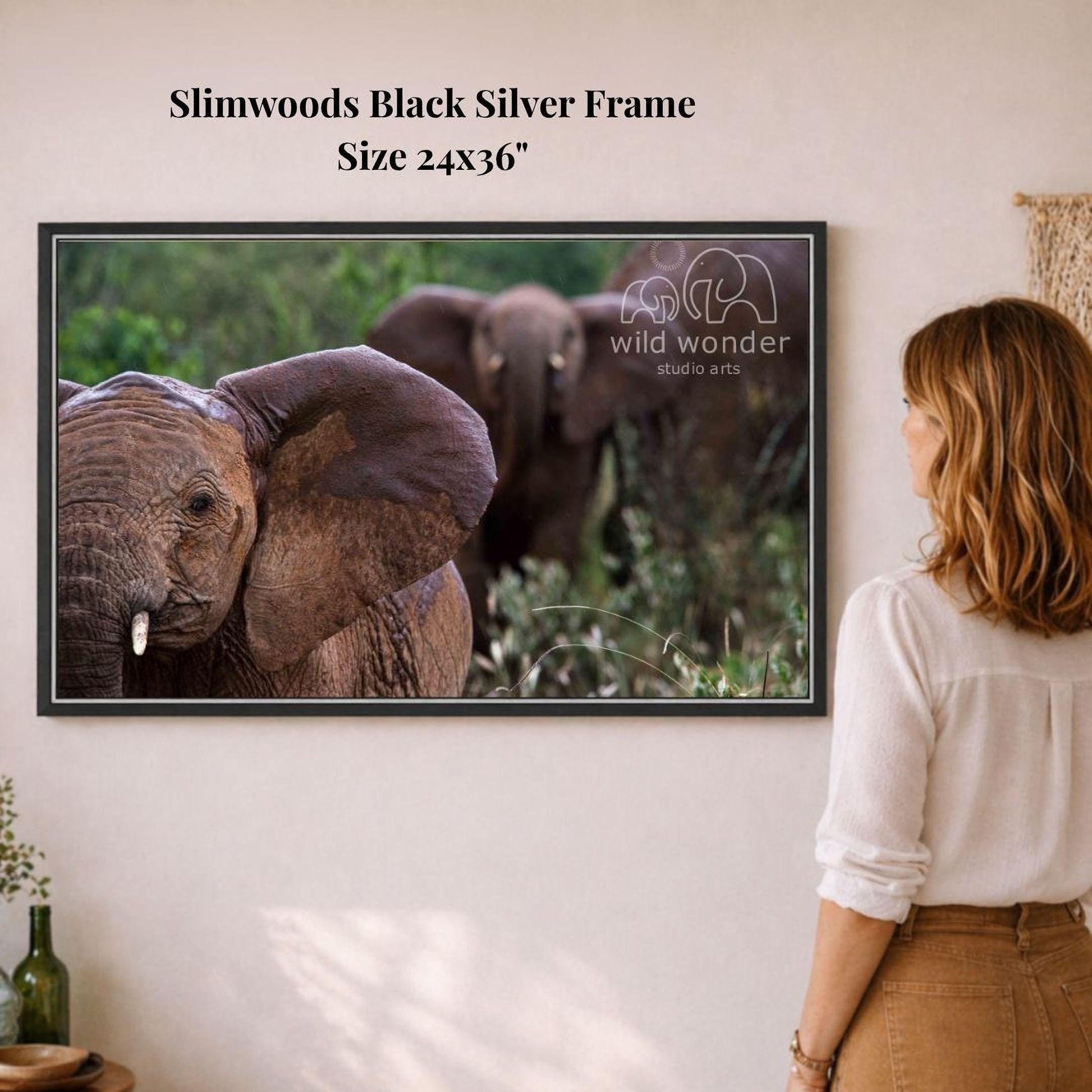 A woman is standing in front of a framed photograph of three elephants in a natural setting, looking at the elephants on the wall.