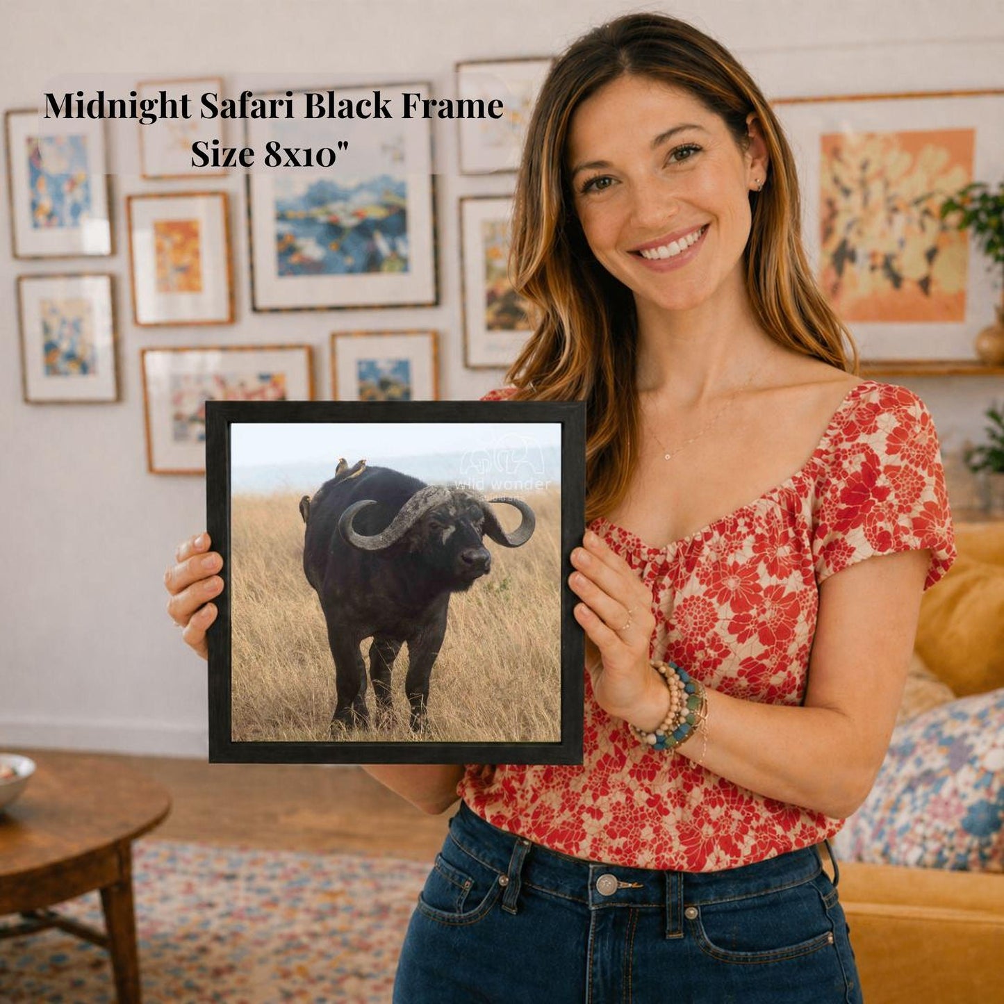 A woman is holding a framed picture of a black ox with a smile on her face, standing in a room with various framed pictures on the wall behind her.